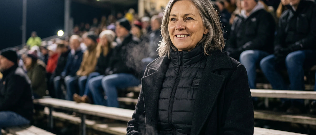 A woman smiling on stadium bleachers at night wearing the black heated vest under her coat